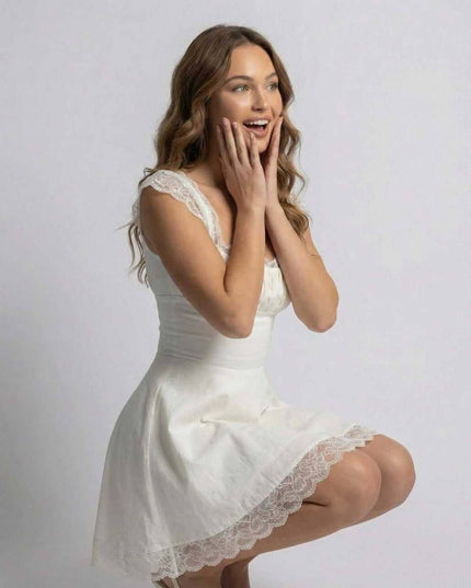 White lace mini dress with square neckline and scalloped hem, worn in a seated pose