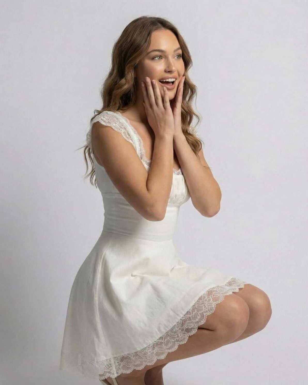 White lace mini dress with square neckline and scalloped hem, worn in a seated pose
