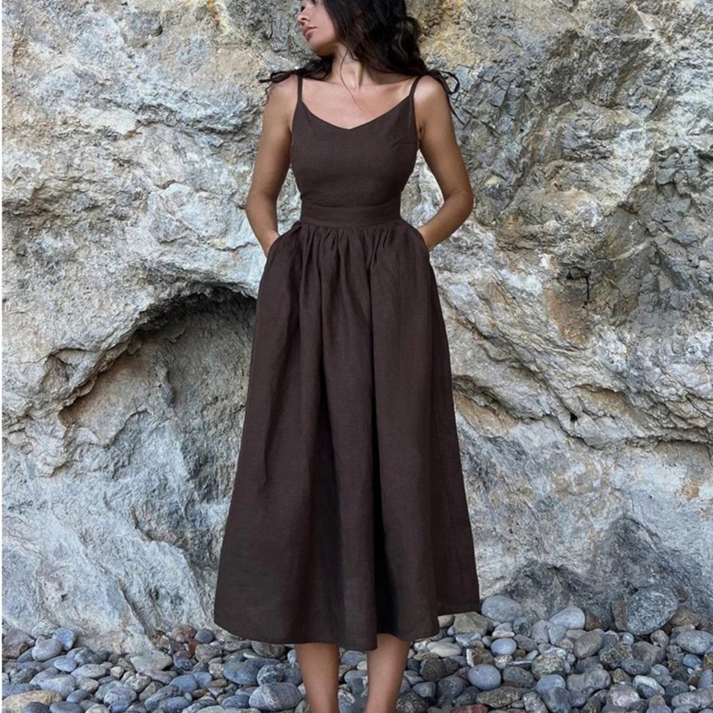 Dark brown linen maxi dress with spaghetti straps and full gathered skirt against rock wall