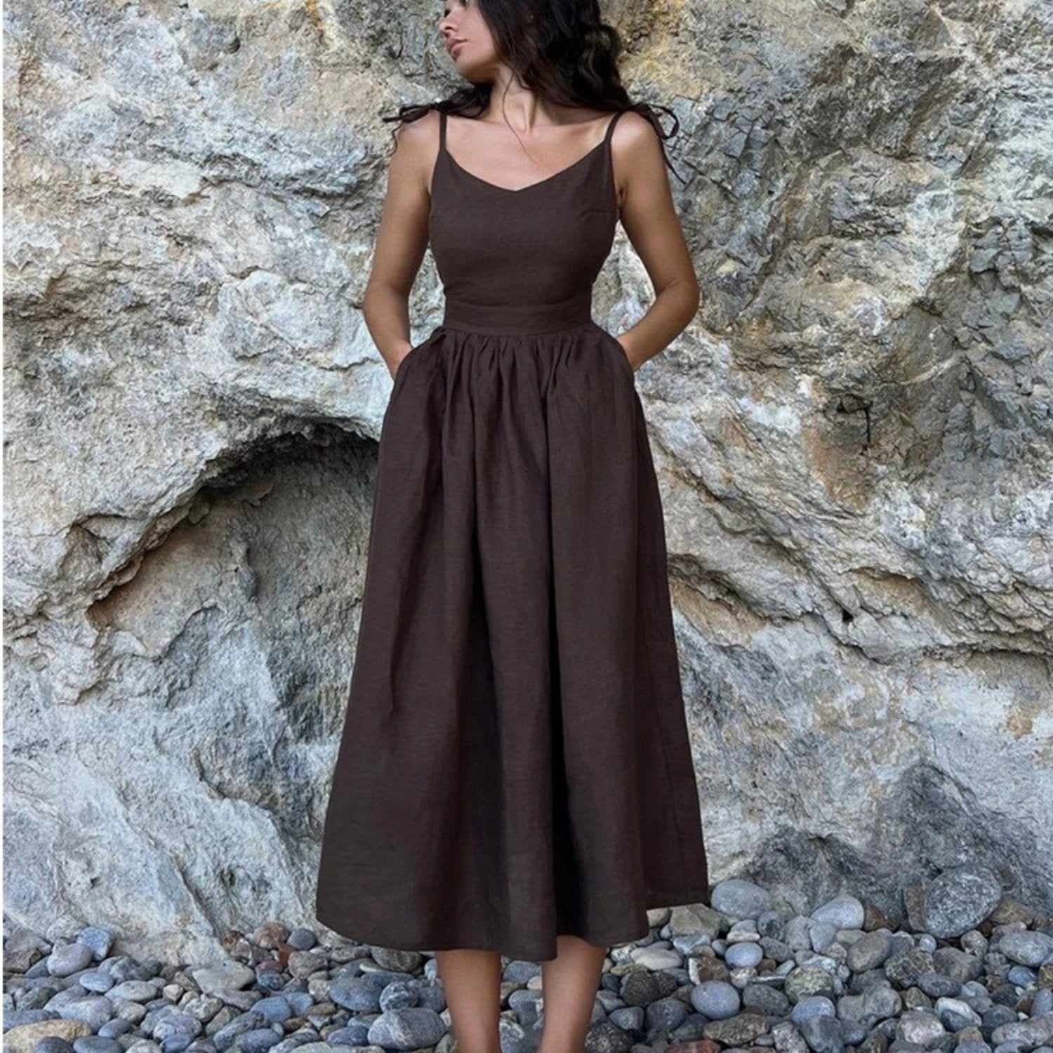 Dark brown linen maxi dress with spaghetti straps and full gathered skirt against rock wall