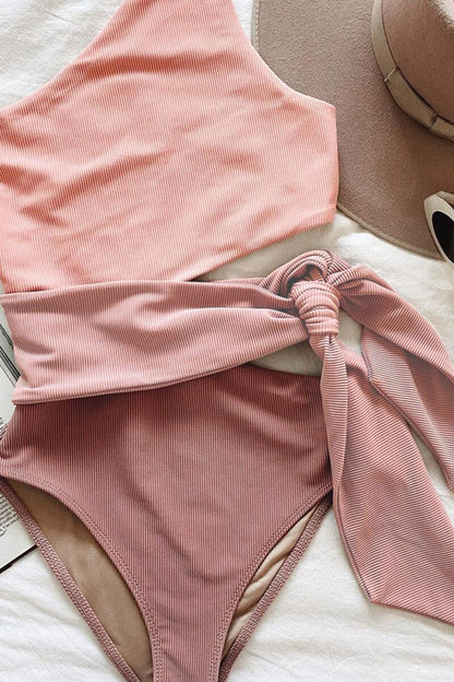 Folded pink ribbed one piece swimsuit with one shoulder cutout and matching waist tie