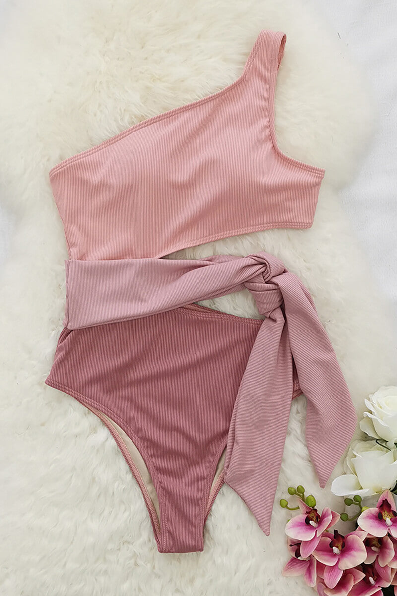 Flat lay of pink one shoulder one piece swimsuit with cutout waist tie on white fur