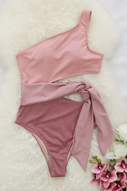 Flat lay of pink one shoulder one piece swimsuit with cutout waist tie on white fur